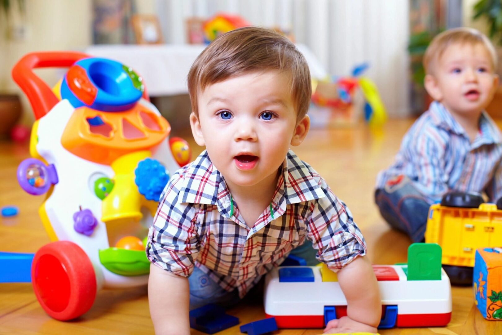 Two toddlers play on the floor with colorful toys. One toddler is in the foreground, looking forward, while the other is in the background.