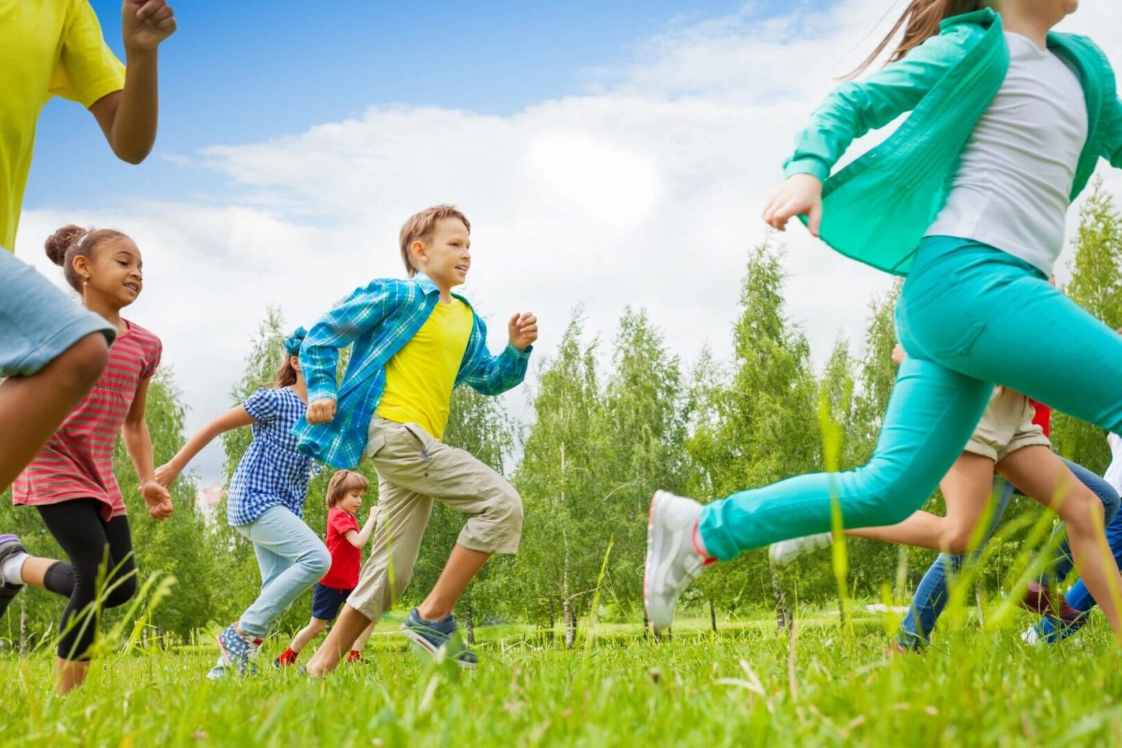 Children running and playing on a grassy field with trees in the background, under a blue sky.