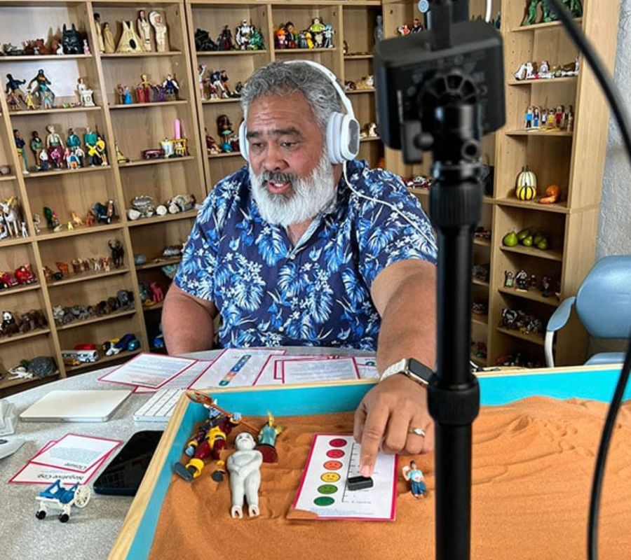 Man painting small figurines at a cluttered desk with shelves full of collectibles.