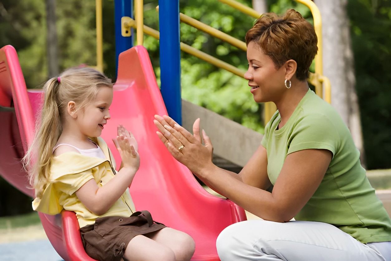A woman and child playing pat-a-cake near a playground slide.