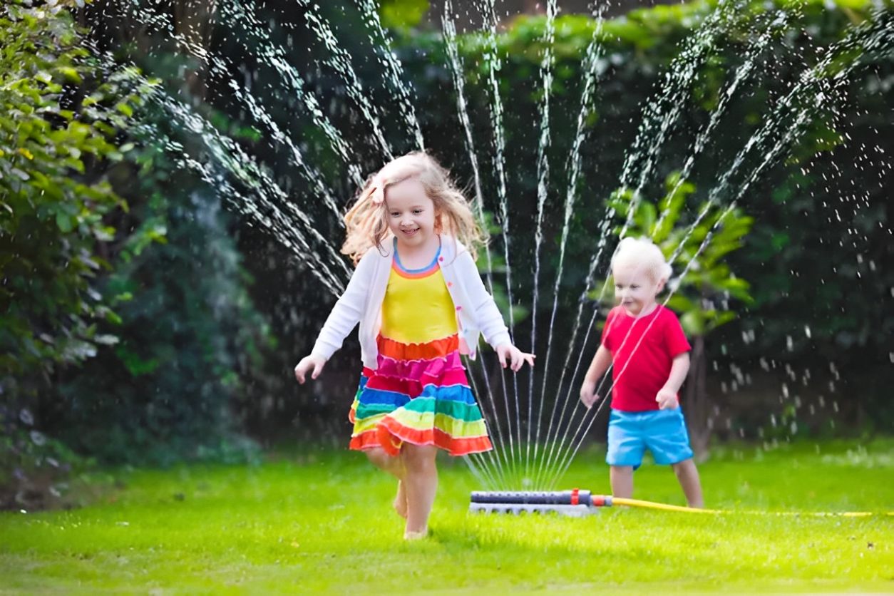 Two children joyfully play in the garden sprinkler on a sunny day.