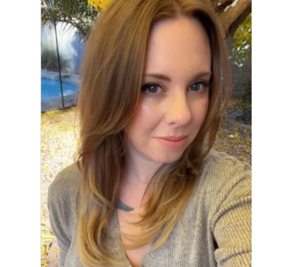 A woman with light brown hair smiling softly in a cozy indoor setting.
