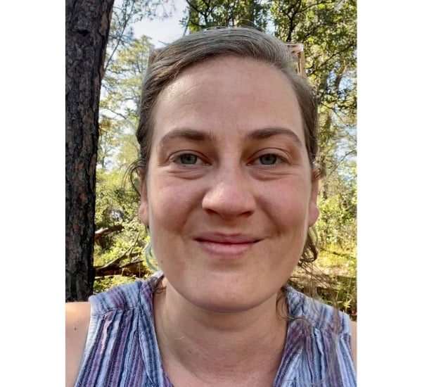 A woman smiling outdoors with trees in the background.