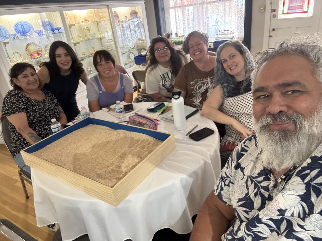 A group of six people smiling around a table with a large cake in a bright room.