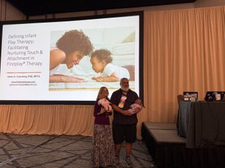Two women on stage holding a baby with a presentation slide behind them.