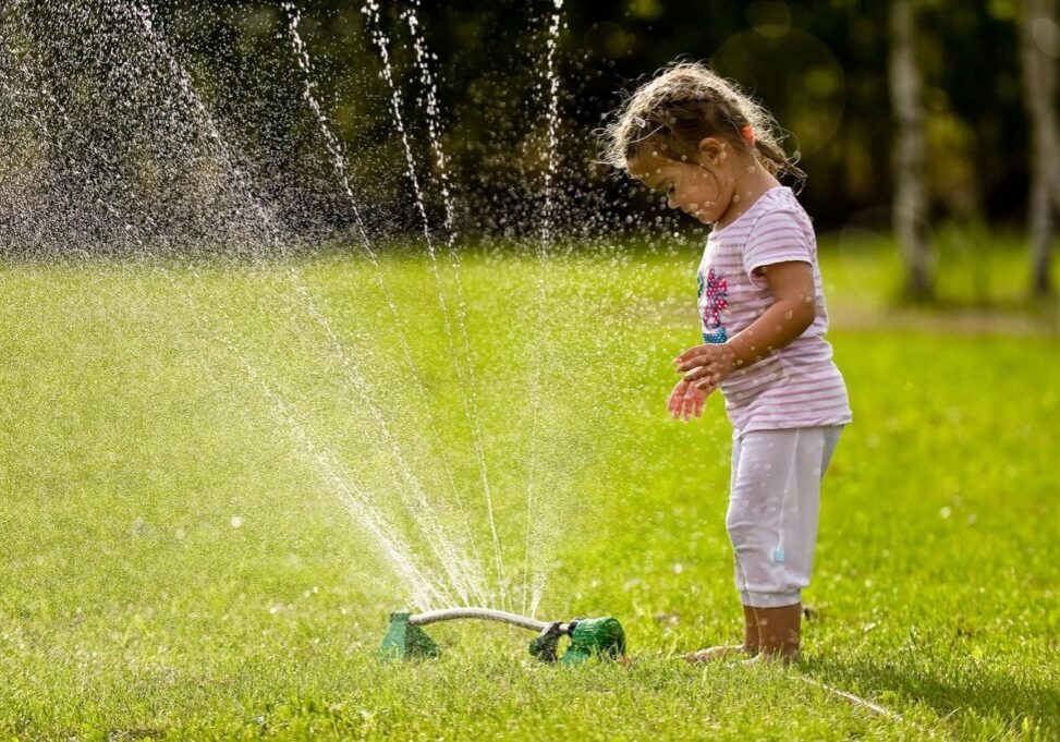 Child in white and pink outfit plays with a garden sprinkler on a sunny day, surrounded by green grass.